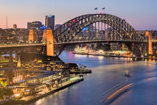 Color photograph of Circular Quay with arcing ferry light trails mirroring Sydney Harbour Bridge from office tower above