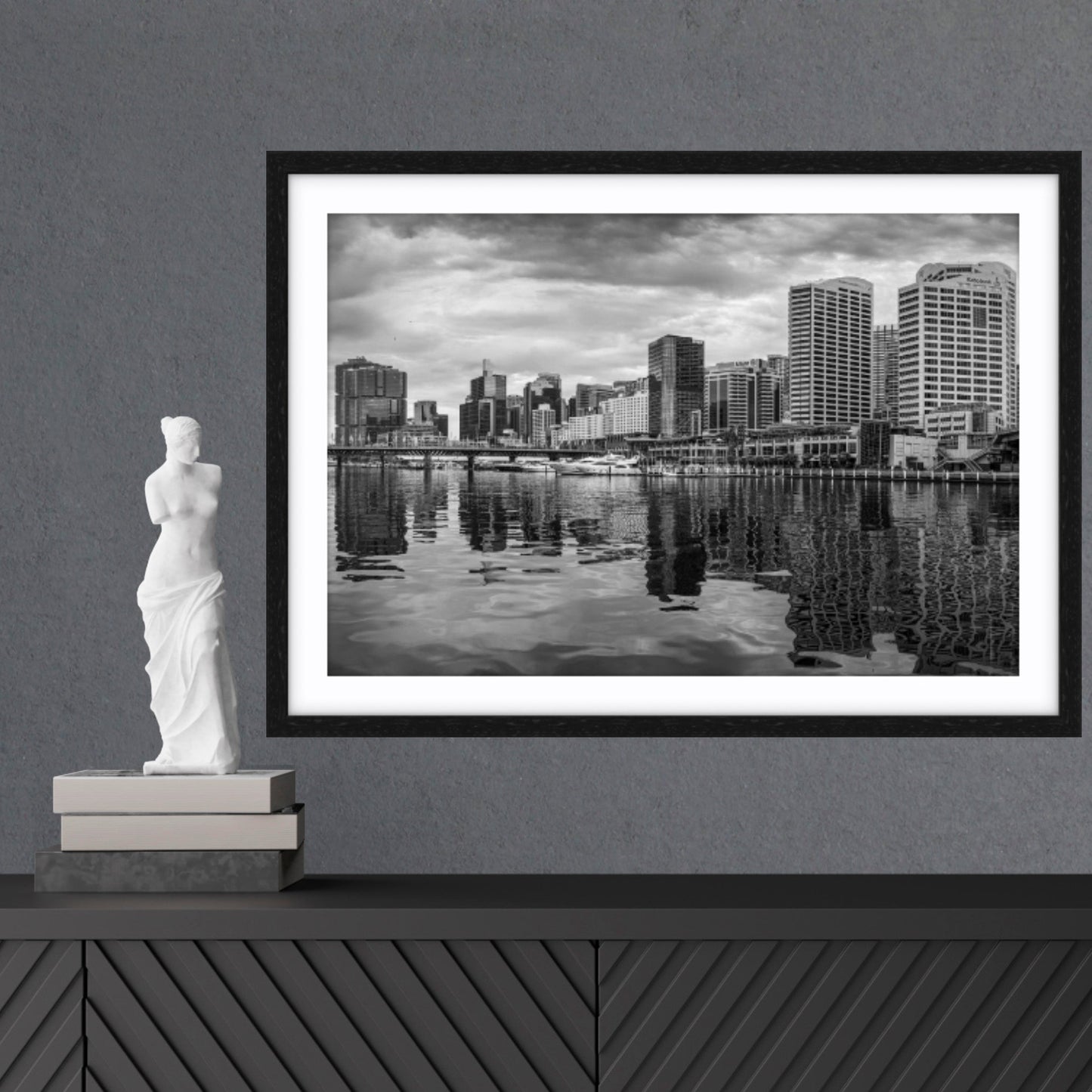 Framed fine art black and white cityscape print on a wall with a statue and books below.
